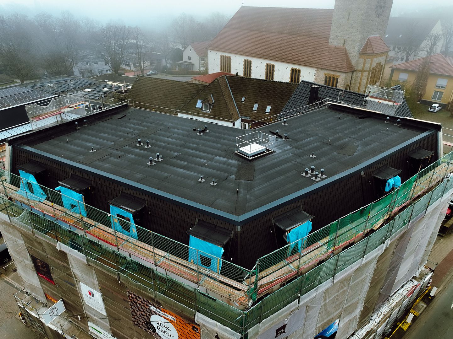 Luftaufnahme eines modernen Flachdachs während der Dachsanierung mit Gerüst an einem Gebäude neben einer Kirche